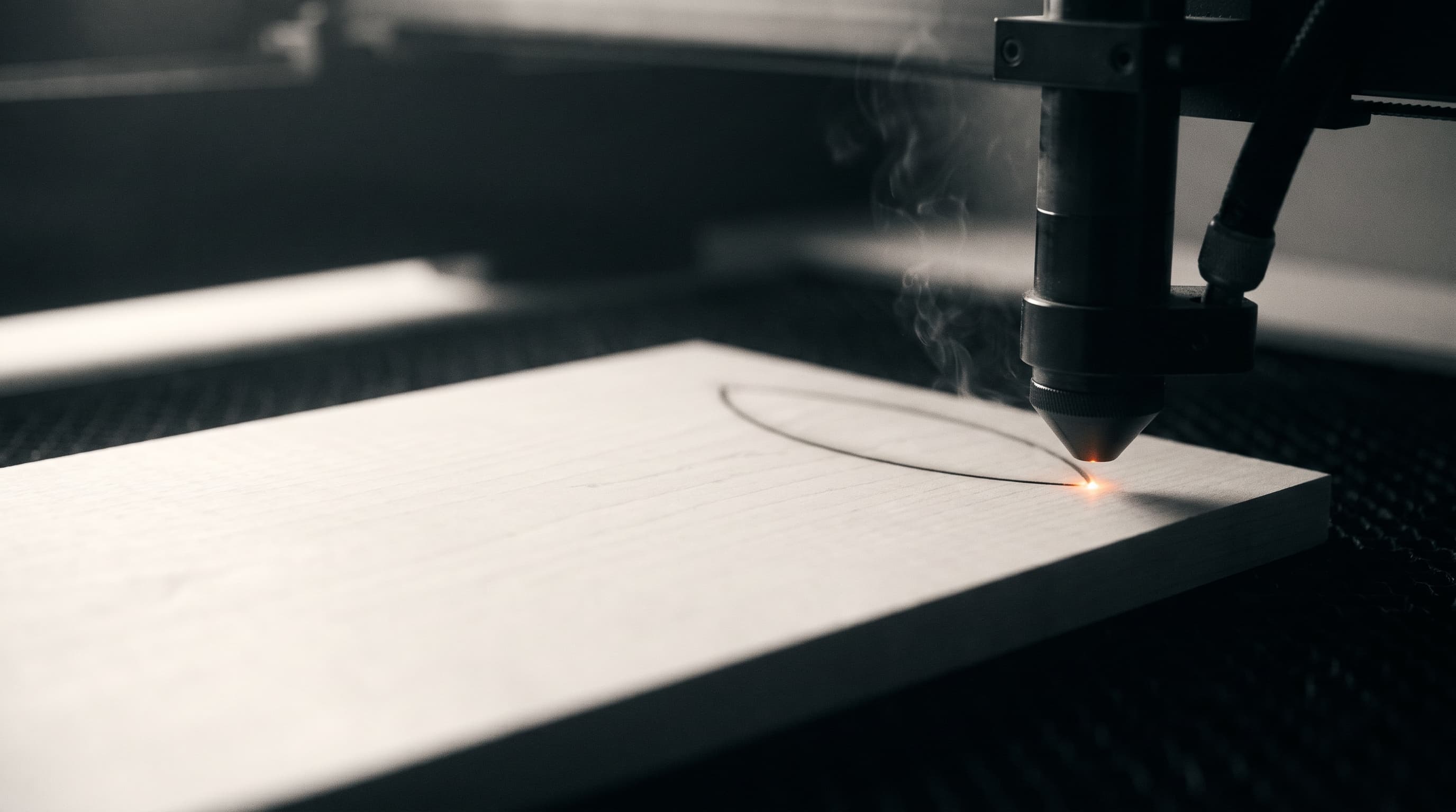 Laserdüse dicht über einer hellen Holzplatte, an der Spitze glüht der Laserpunkt, darunter eine gebrannte Linie
